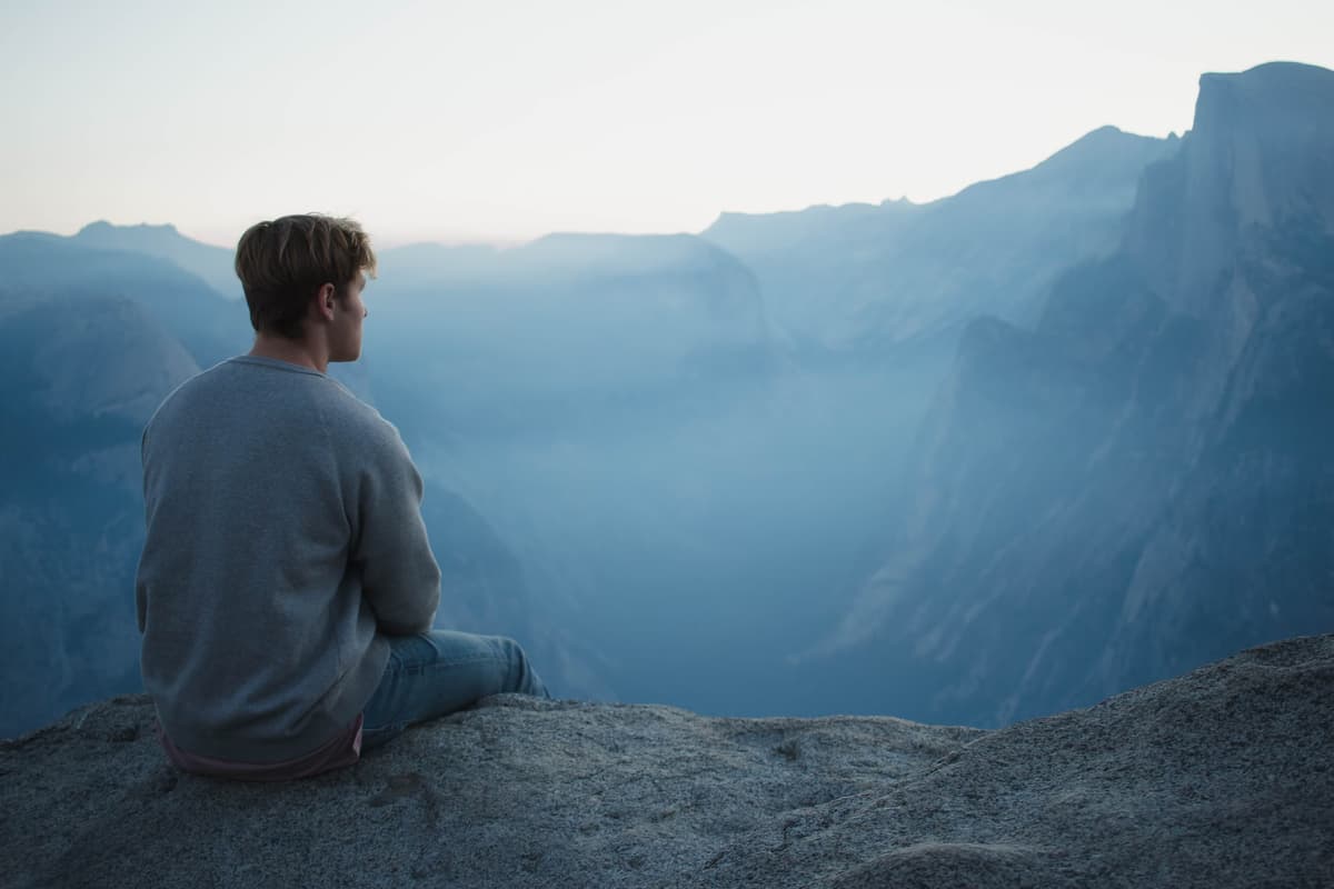 Un homme assis seul face à des montagnes bleutées, dans un moment de calme propice à la méditation